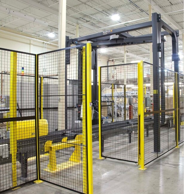 Manufacturing cell enclosed by black mesh safety fencing with yellow posts in a factory, containing industrial equipment and a conveyor system.