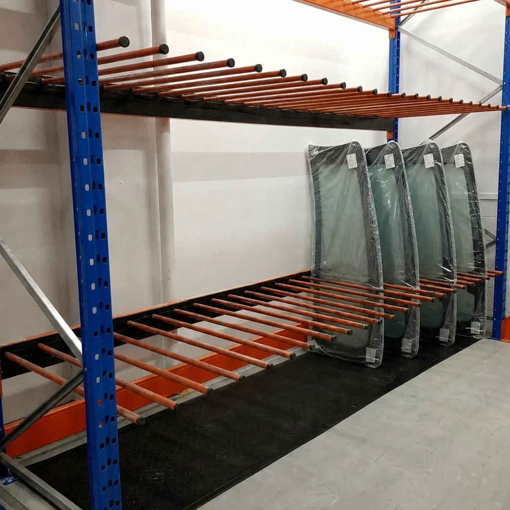 Rangement vertical en industrie automobile equipement entreposage stockage Pedlex fabrique au Quebec - Pedlex Blue metal warehouse rack with orange pipe-like supports on two levels; several wrapped mattresses standing against the rack on the right side.